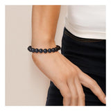Une personne portant le Bracelet NOELYSE de Perline - un bracelet en Argent & Perles Noires 9/10 mm avec un mousqueton en fermoir - au poignet, associé à un top blanc et un pantalon sombre, la main détendue sur le côté.