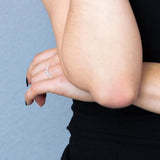 Une personne en noir croise les bras, mettant en valeur la Bague Solitaire | Argent | Enveloppée, ornée d'oxydes de zirconium blancs. Ses ongles noirs contrastent avec un simple mur bleu clair.